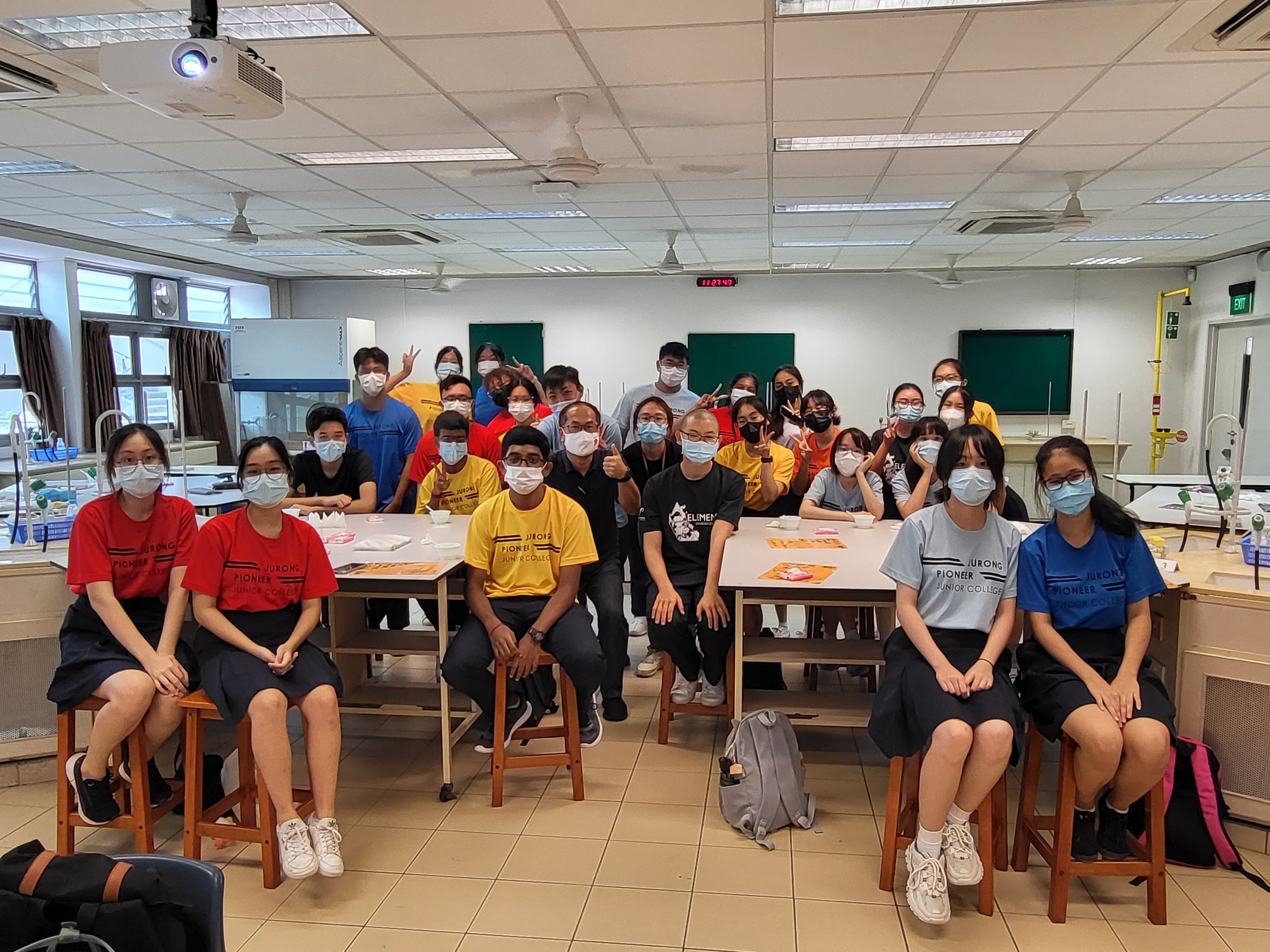 A large group of students and teachers wearing face masks pose together in a school laboratory classroom.
